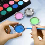 Hand holding a brush dipping into a stainless steel paint mixing tray filled with blue paint, with other trays containing pink and green paint in the background.